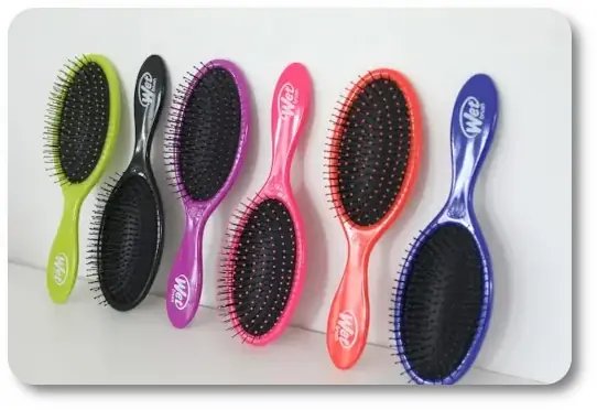 Row of colorful Wet Brushes lined up against a white wall, featuring green, black, pink, orange, and purple handles with black bristles.